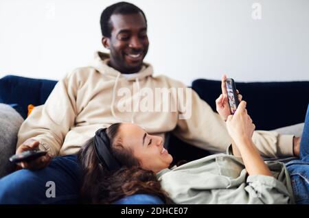 Junge gemischte Ethnizität Paar Entspannung auf dem Sofa zu Hause mit Mann beim Fernsehen, während Frau auf dem Schoß liegt und kontrolliert Soziale Medien auf dem Mobiltelefon Stockfoto