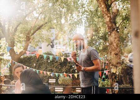 Junger Mann mit rasierten Kopf, der im Stehen durch Kopfhörer spricht Auf dem Balkon während der Party Stockfoto