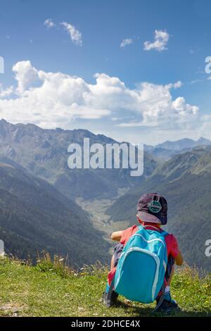 Abenteuerlicher und unerschrockener kleiner Junge mit blauer Mütze und Rucksack, der auf dem Rücken hockt, beobachtet von der Spitze eines Berges das Tal eines Berges Stockfoto