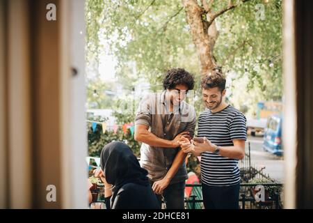 Glücklicher junger Mann zeigt Handy zu männlichen Freund, während Während der Party auf dem Balkon stehen Stockfoto