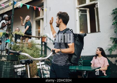 Lächelnder junger Mann winkt Hand, während er mit dem Fahrrad steht Hinterhof Stockfoto