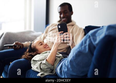 Junge gemischte Ethnizität Paar Entspannung auf dem Sofa zu Hause mit Mann beim Fernsehen, während Frau auf dem Schoß liegt und kontrolliert Soziale Medien auf dem Mobiltelefon Stockfoto