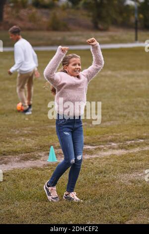 Lächelnde Schwester mit erhobenen Armen stehen auf grasbewachsenen Feld, während Bruder spielt Fußball im Hintergrund im Park Stockfoto