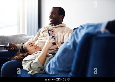 Junge gemischte Ethnizität Paar Entspannung auf dem Sofa zu Hause mit Mann beim Fernsehen, während Frau auf dem Schoß liegt und kontrolliert Soziale Medien auf dem Mobiltelefon Stockfoto