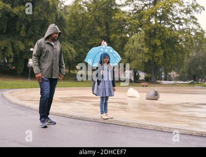 Volle Länge des Vaters Blick auf Tochter mit Regenschirm, während Stehen im Park bei Regenfällen Stockfoto