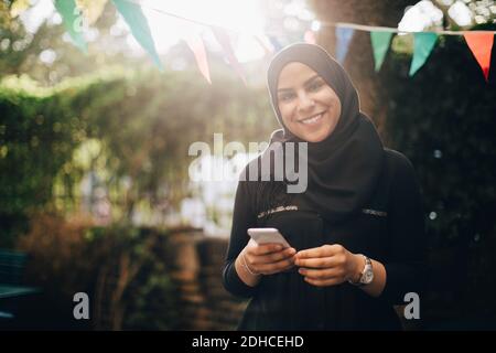 Porträt einer lächelnden Frau in Hijab hält Smartphone an Hinterhof Stockfoto