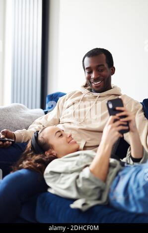 Junge gemischte Ethnizität Paar Entspannung auf dem Sofa zu Hause mit Mann beim Fernsehen, während Frau auf dem Schoß liegt und kontrolliert Soziale Medien auf dem Mobiltelefon Stockfoto
