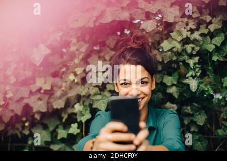 Porträt einer lächelnden jungen Frau, die über das Mobiltelefon fotografiert Hinterhof Stockfoto