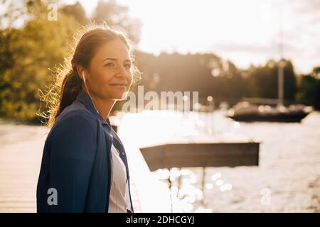 Nachdenkliche Frau hört Musik über Kopfhörer, während sie am See sitzt Im Sommer Urlaub Stockfoto