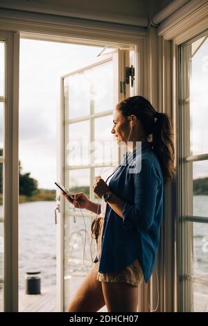 Durchdachte lächelnde Frau, die während des Gesprächs über Kopfhörer auf dem Smartphone spricht Stehen an der Ferienvilla Stockfoto