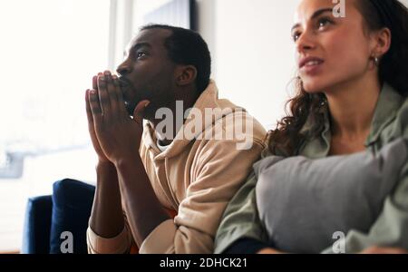 Aufgeregt und angespannt junge gemischte Ethnizität Paar auf dem Sofa sitzen Zu Hause Sportereignisse im fernsehen anschauen oder streamen Stockfoto