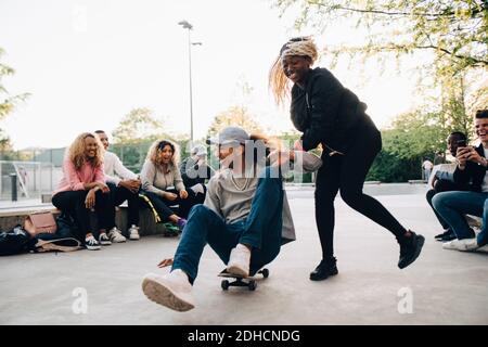 Teenager-Mädchen schieben jungen Mann auf Skateboard, während Freunde lachen Im Park Stockfoto