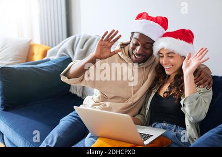 Junge gemischte ethnische Zugehörigkeit Paar trägt Santa Hüte entspannen auf Sofa Zu Hause machen Weihnachten Videoanruf auf Laptop zur Familie Und Freunden Stockfoto