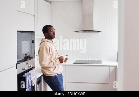 Unglücklicher junger Mann mit psychischen Problemen stehen in der Küche Zu Hause mit heißem Getränk Stockfoto