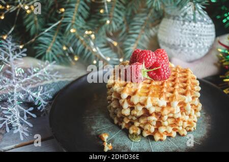 Wiener Waffeln mit Himbeeren und Honig auf dem Weihnachtstisch mit Christbaumzweigen und Girlande-Lichtern im Hintergrund. Stockfoto