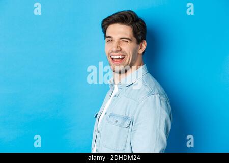 Glücklicher und selbstbewusster Mann dreht den Kopf an die Kamera, zwinkend und lächelnd, stehend über blauem Hintergrund Stockfoto