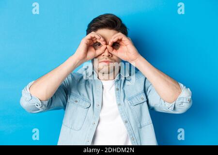 Nahaufnahme eines Mannes, der die Hände auf die Augen hält, durch ein Fernglas mit ernstem Gesicht blickt und auf blauem Hintergrund steht Stockfoto