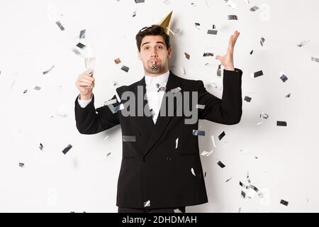 Schöner Party-Kerl im schwarzen Anzug Spaß haben, feiern Neujahr, weht Pfeife und trinken Champagner, während Konfetti fallen Stockfoto