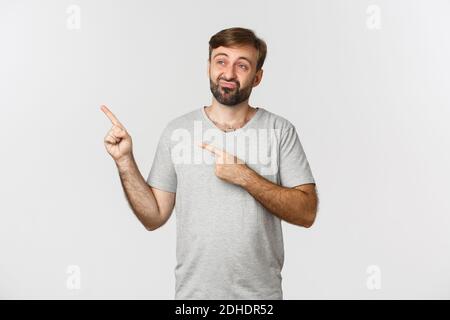 Bild von skeptischem und unamüsiertem bärtigen Kerl in grauem T-Shirt, Pouting und Zeigefinger auf die obere linke Ecke, über wh stehen Stockfoto