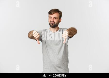 Porträt eines skeptischen bärtigen Mannes im lässigen T-Shirt, zeigt Daumen nach unten und sieht enttäuscht, missbilligen etwas Schlechtes, sta Stockfoto