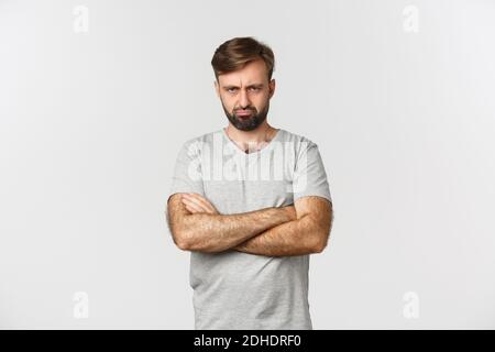 Porträt von beleidigt gut aussehenden Mann, schmollend und wütend Grimasse, Kreuz Hände über Brust defensiv, stehend über weißen Rücken Stockfoto