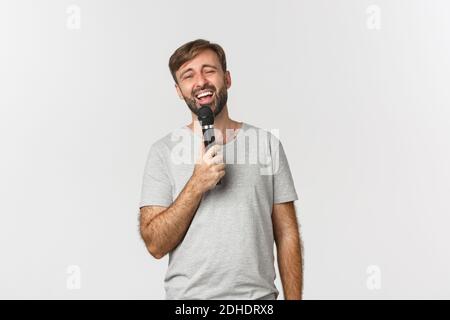 Schöner sorgloser Kerl, der Lied in Karaoke singt, Mikrofon hält und auf weißem Hintergrund steht Stockfoto