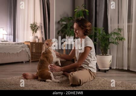 Fröhliche junge Frau lächelt und spielt mit Hund während der Ruhe Auf dem Boden zu Hause Stockfoto