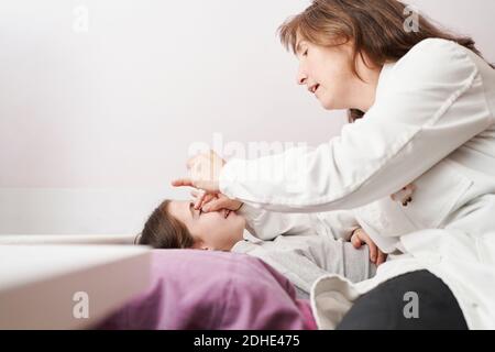 Ärztin lächelt und gibt einem kleinen Mädchen in ihrem Bett Augentropfen. Hausarzt Konzept Stockfoto