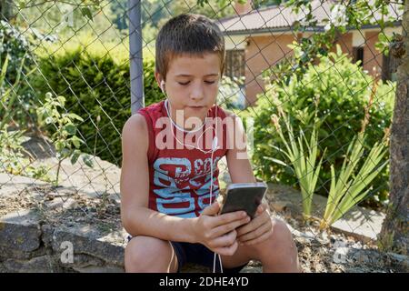 Junge, die ihre Smartphones betrachten und Musik hören, mit Kopfhörern, die an sonnigen Tagen auf einer Steinwand sitzen. Technologiekonzept Stockfoto