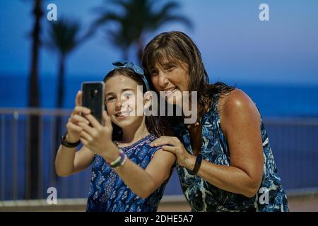 Porträt von Mutter und Tochter im Teenageralter in blauen Kleidern und machen ein Selfie mit ihrem Smartphone in der Nacht mit einem Strand im Hintergrund. Vacati Stockfoto
