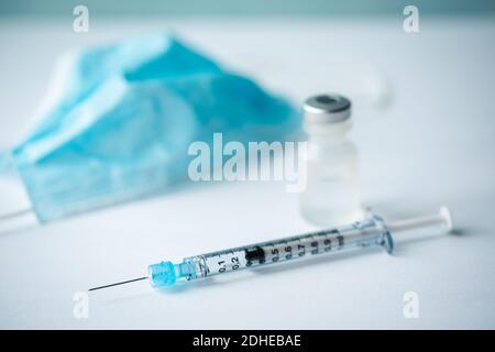Nahaufnahme von Nadel, Spritze, Maske und Fläschchen mit Impfstoff auf weißem Tisch. Stockfoto