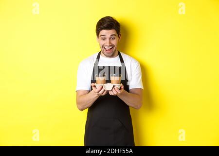Der Kellner schaute aufgeregt auf zwei Tassen Kaffee zum Mitnehmen, trug eine schwarze Schürze und stand auf gelbem Hintergrund Stockfoto