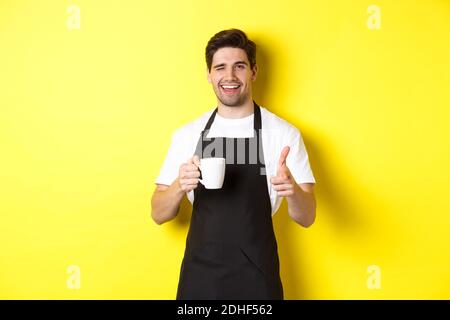 Ein hübscher Barista in schwarzer Schürze hält eine Kaffeetasse, zeigt mit dem Finger auf dich und lädt dazu ein, Café zu besuchen, das auf gelbem Hintergrund steht Stockfoto