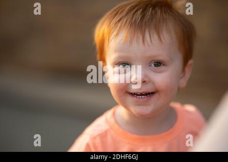 Ein entzückender, rothaariger kleiner Junge in einem orangefarbenen Hemd (mein Sohn) lächelt bei einem warmen Licht am späten Nachmittag an der Kamera. Stockfoto