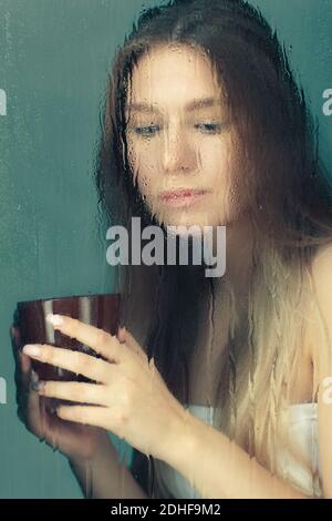 Porträt einer jungen blonden Frau mit Kaffeetasse in den Händen hinter dem Fenster Glas mit Regentropfen. Stockfoto