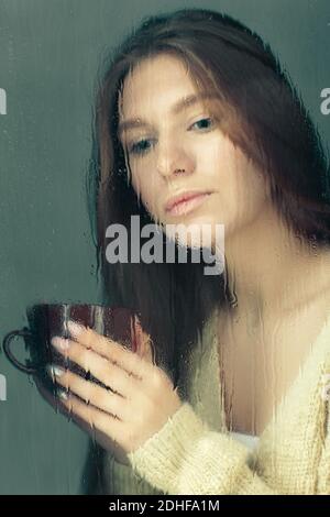 Porträt einer jungen blonden Frau mit Kaffeetasse in den Händen hinter dem Fenster Glas mit Regentropfen. Stockfoto