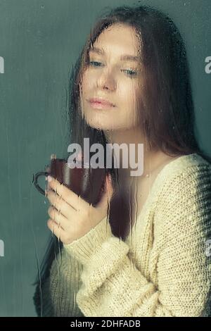 Porträt einer jungen blonden Frau mit Kaffeetasse in den Händen hinter dem Fenster Glas mit Regentropfen. Stockfoto