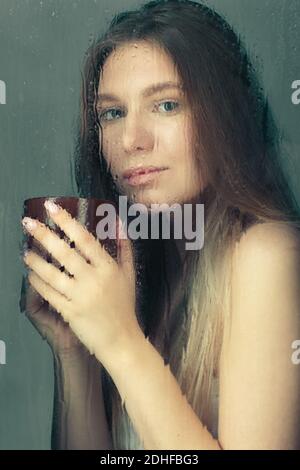 Porträt einer jungen blonden Frau mit Kaffeetasse in den Händen hinter dem Fenster Glas mit Regentropfen. Stockfoto