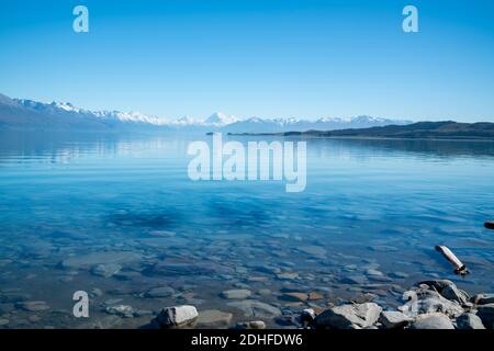 Küstenlinie und Blick über den ruhigen Pukaki-See mit Mount Cook und den südlichen Alpen in der Ferne. Stockfoto