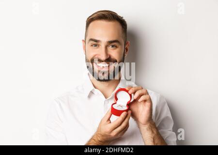 Nahaufnahme des glücklichen, gutaussehenden Mannes, der einen Vorschlag macht, den Ehering in der Box hält und lächelt, ihn bittet, ihn zu heiraten, weißer Hintergrund Stockfoto