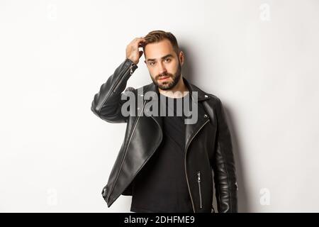 Schöner Macho-Mann in schwarzer Biker-Jacke, berührt seine Frisur und sieht cool, weißen Hintergrund Stockfoto