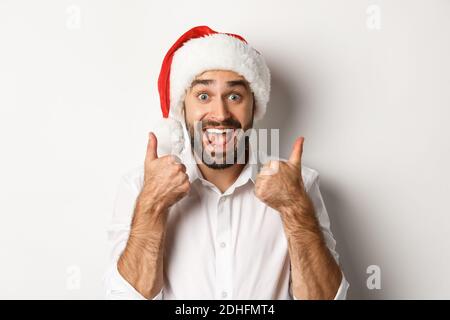 Party, Winterferien und Feiertagskonzept. Mann genießt weihnachten, trägt weihnachtsmütze und zeigt Daumen mit aufgeregt Gesicht Stockfoto