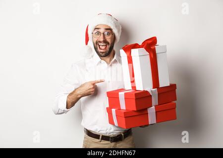 Frohe weihnachten, Urlaub Konzept. Überrascht Mann empfangen Weihnachtsgeschenke, zeigt auf Geschenke und lächelt glücklich, trägt weihnachtsmütze, Stockfoto