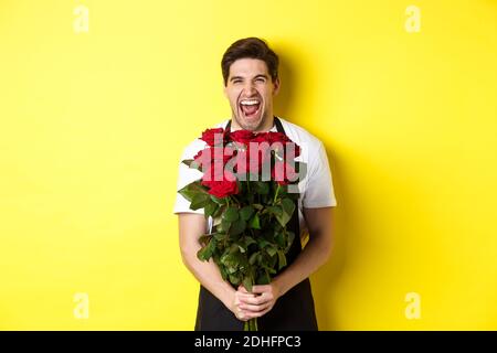 Lustiger junger Verkäufer in schwarzer Schürze, der Strauß Rosen hält, Blumenhändler lachend und über gelbem Hintergrund stehend Stockfoto