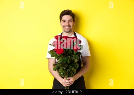 Lächelnder Florist in schwarzer Schürze, der Blumen hält, Rosenstrauß verkauft, der über gelbem Hintergrund steht Stockfoto