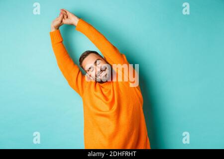 Bild eines hübschen jungen Mannes, der sich die Hände streckt und nach guter Ruhe lächelt und in einem orangefarbenen Pullover auf hellblauem Hintergrund steht Stockfoto