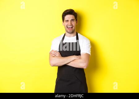 Ein hübscher Barista in schwarzer Schürze winkend zu dir, trägt schwarze Schürzenuniform und steht auf gelbem Hintergrund Stockfoto