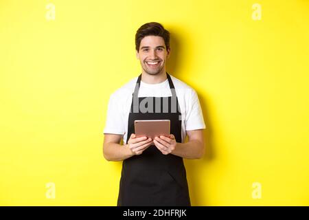 Kellner in schwarzer Schürze nimmt Aufträge entgegen, hält digitale Tablette und lächelt freundlich, steht auf gelbem Hintergrund Stockfoto