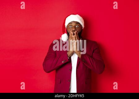 Weihnachts-, Party- und Feiertagskonzept. Schockiert afroamerikanischen Kerl in santa hat Reaktion sprachlos auf Werbung, stehend Stockfoto