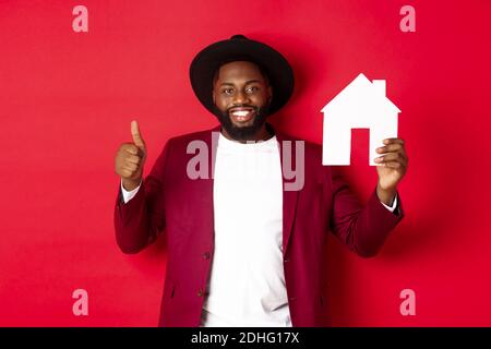 Immobilien. Stilvoller afroamerikanischer bärtiger Mann, der Hausmaket zeigt, Daumen hoch, Agentur oder Apartment-Komplex empfiehlt, Standdi Stockfoto